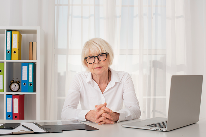 Older female teacher in glasses sitting at desk with laptop, representing a classroom incident involving a student and suspension issue.
