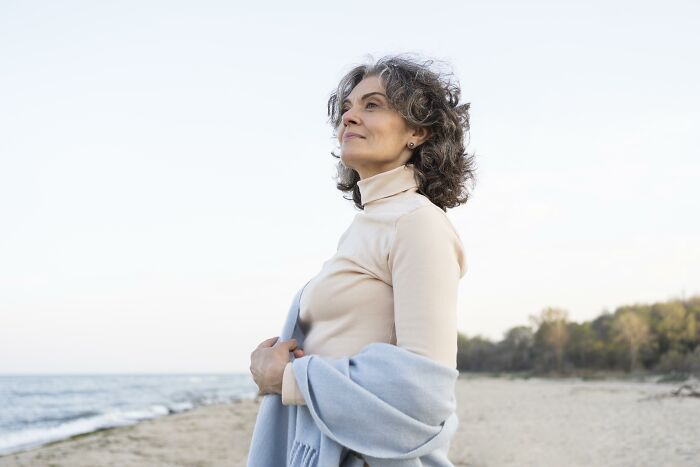 Middle-aged woman standing on beach wrapped in shawl, reflecting on things people would never admit in real life.