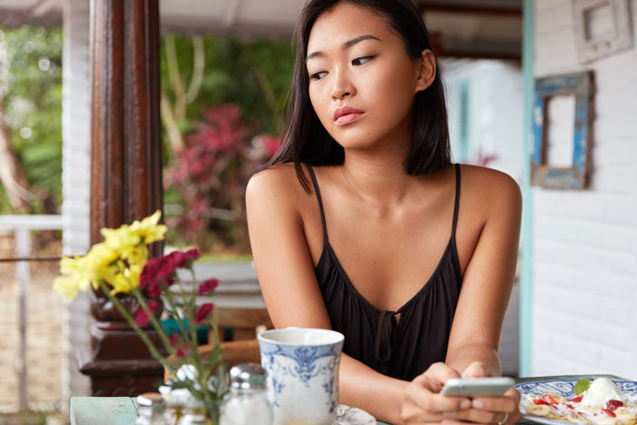 Young woman holding phone at a table with food, appearing thoughtful while considering ordering fried pork intestine.