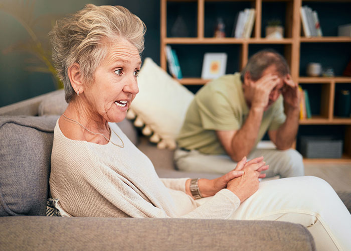Older woman angrily talking while man sits stressed in background, illustrating parents made son raise siblings conflict.