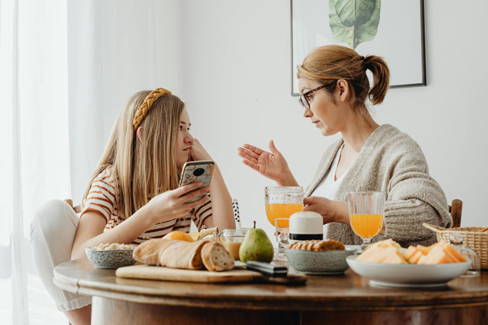 A 19-year-old girl looks irked while talking to a woman over breakfast, reflecting forced parenting of little sister.
