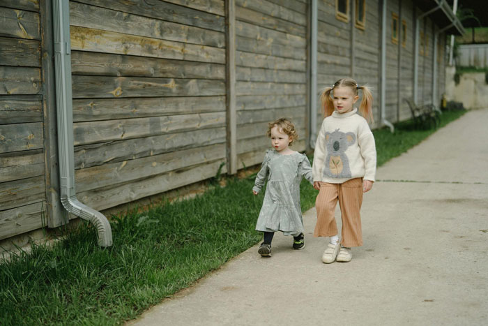 Two young sisters walking outside along a wooden fence, depicting forced parenting of little sister by 19YO sibling.