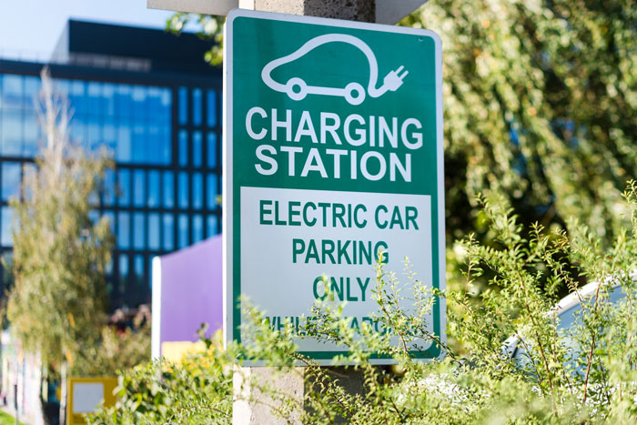 Green electric car charging station sign indicating parking for EVs only near a building with trees and bushes around.