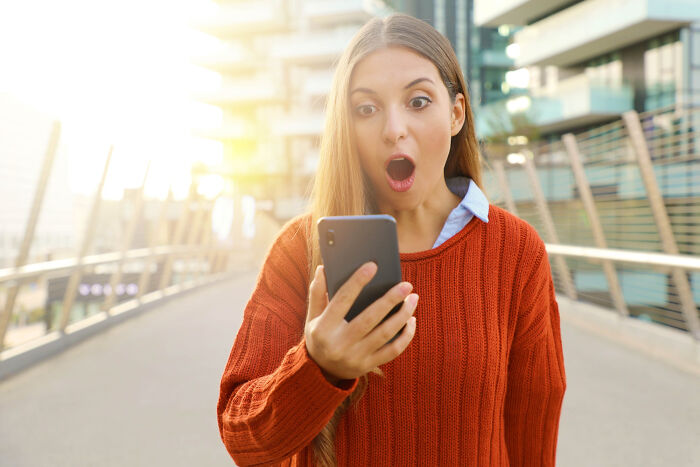 Young woman in orange sweater looking at partner's phone with shocked expression outdoors.