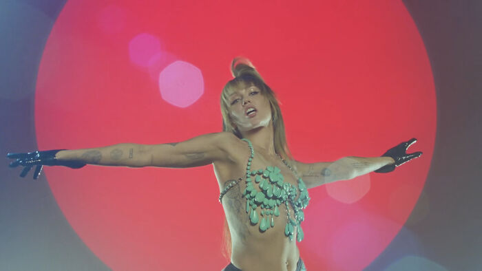 Performer in a bold outfit with turquoise details and black gloves posing with arms outstretched against a red circular background.