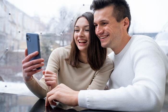 Couple smiling and taking a selfie together using a phone, enjoying a casual moment inside a cozy café.