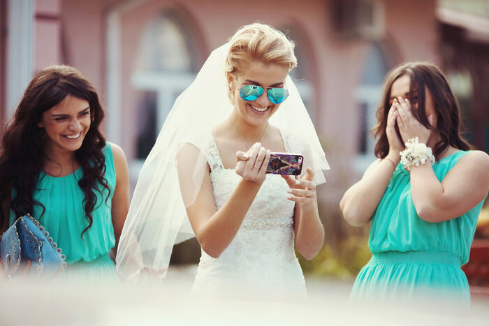 Bride smiling at partner's phone while bridesmaids react with surprise and joy outdoors