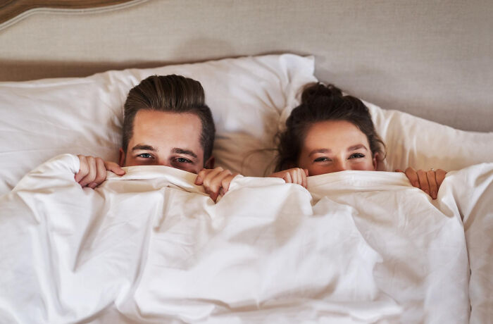 Couple smiling and hiding under white blankets in bed, sharing a playful and cozy moment together.