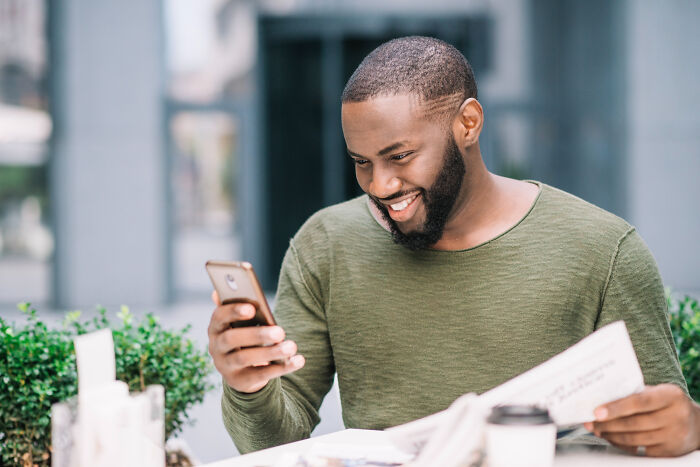 Man smiling while checking partner's phone outdoors in casual green shirt.