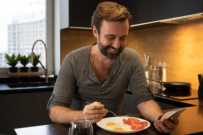 Man smiling while using partner's phone, eating breakfast with eggs and tomatoes in modern kitchen.