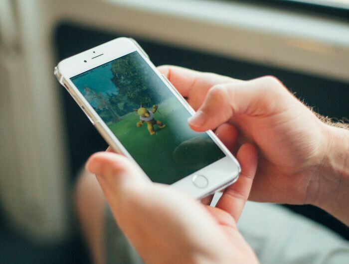 Person holding partner's phone playing an augmented reality game with cartoon character outdoors on the screen.