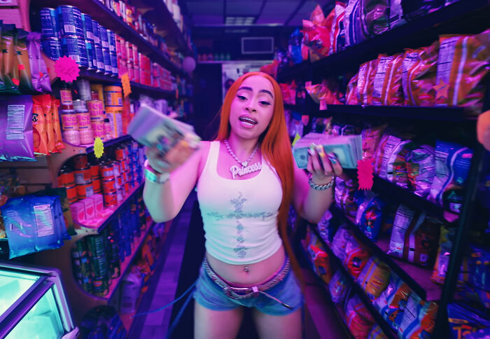 Young woman with red hair holding cash in a colorful convenience store aisle.