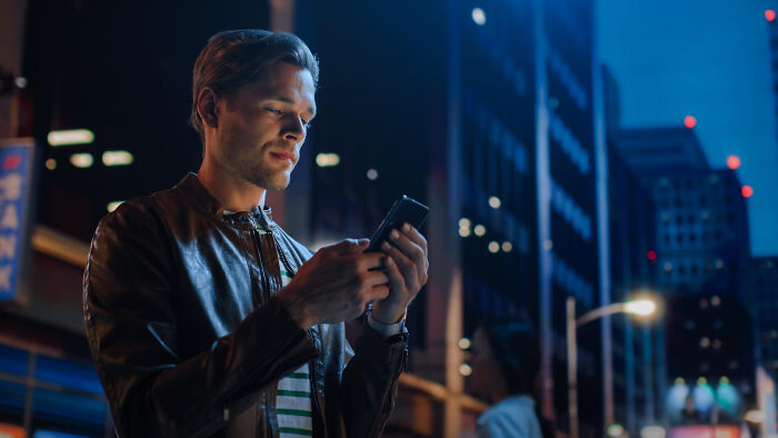 Young man using partner's phone on a city street at night with buildings and streetlights in the background.