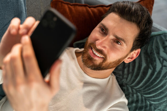 Man lying on couch, smiling while holding partner's phone, relaxing in cozy home environment.