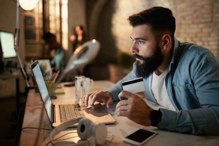 Man using laptop and holding credit card for online payment in a dimly lit workspace.