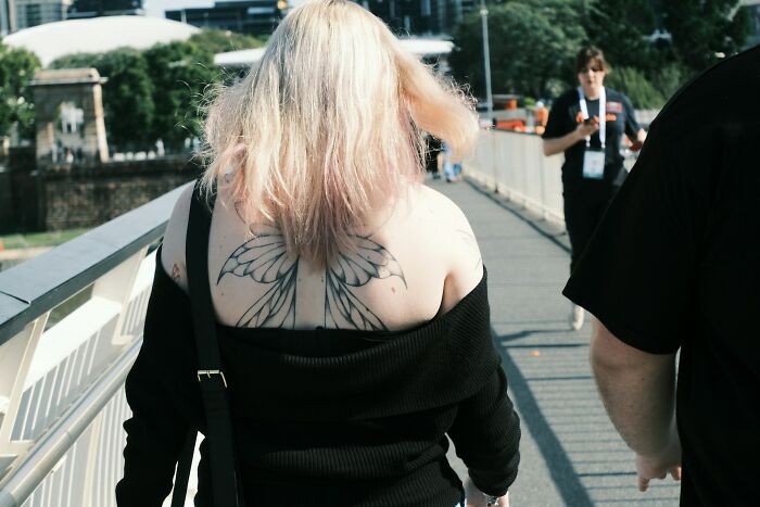 Person with blonde hair and wing tattoos on upper back walking outdoors with others on a sunny day, showcasing tattoo art.