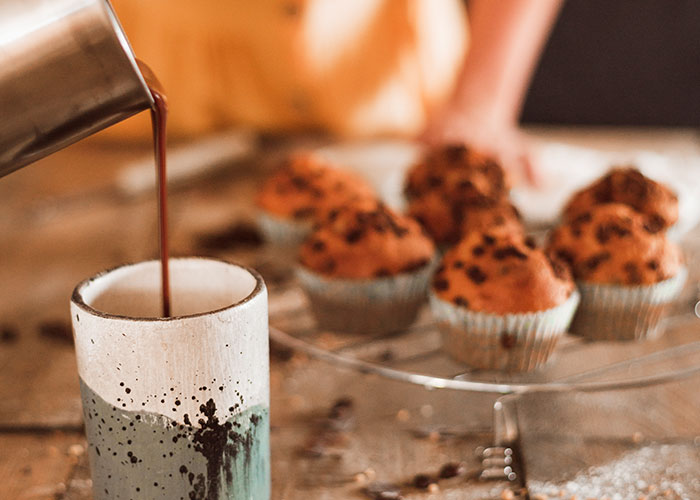Pouring hot chocolate into a ceramic mug with chocolate chip muffins cooling on a wire rack in the background.
