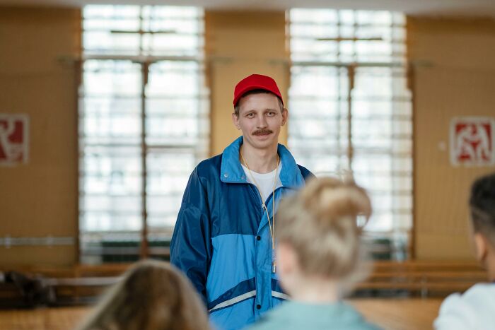Teacher in a red cap and blue jacket delivering funny jokes and iconic comments to surprised students in a classroom setting.