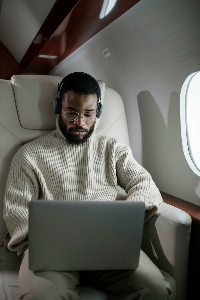 Man wearing headphones and glasses working on a laptop in a private plane, portraying corporate hell escape concept.