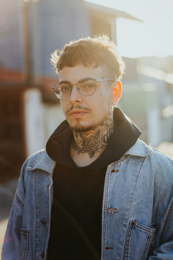 Young tattoo artist with neck tattoos and glasses wearing denim jacket outdoors in natural light, facing the camera.