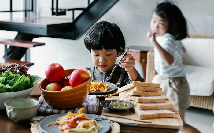 Young child eating lunch at home with another child nearby, illustrating parenting hacks that save sanity.