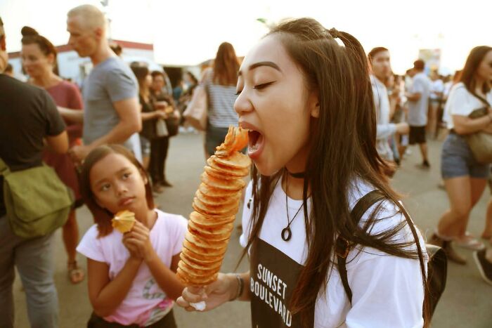 Young woman sharing food at a busy outdoor event, with people around making sounds and looking extremely angry.