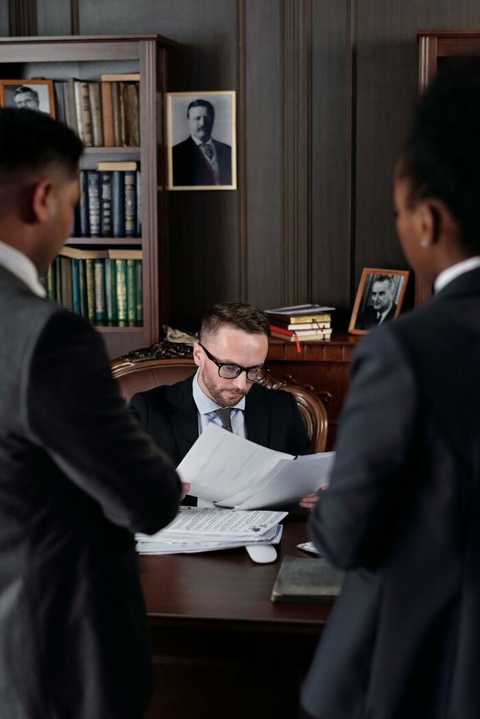 Man in suit reviewing documents at desk while two professionals stand nearby in a vintage office setting for ultra-wealthy jobs