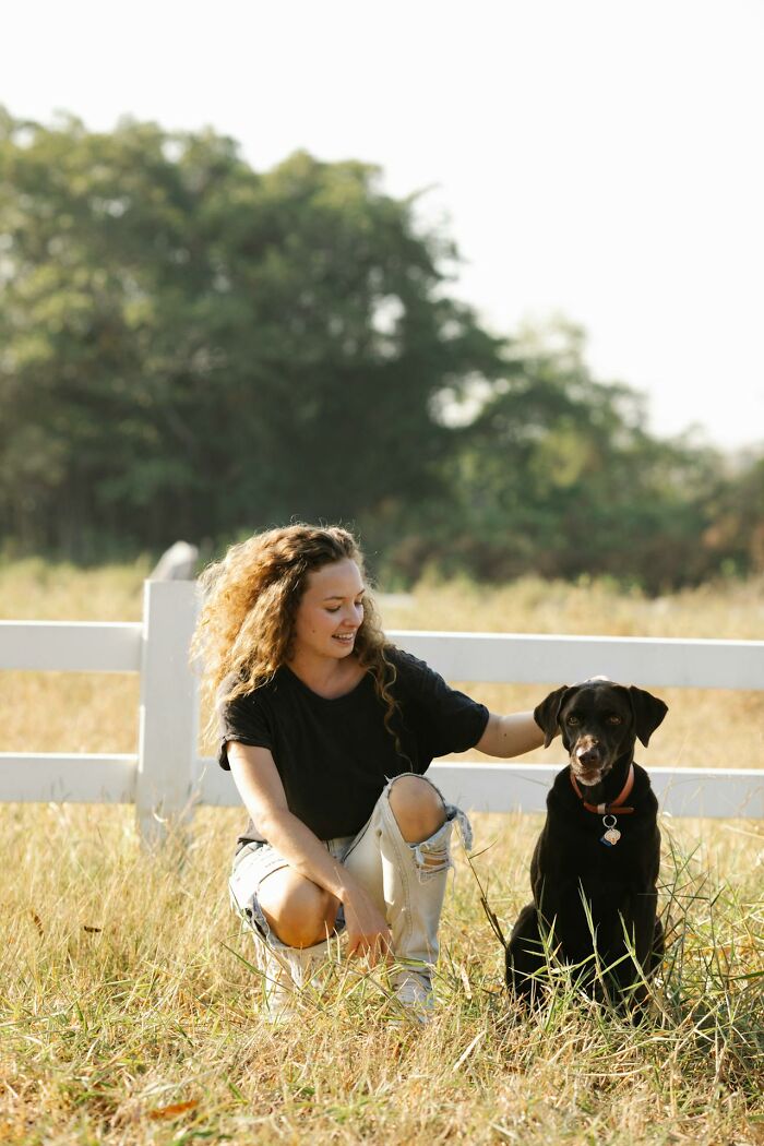 Young woman with curly hair crouching and petting a black dog while alone in the woods near a white fence.