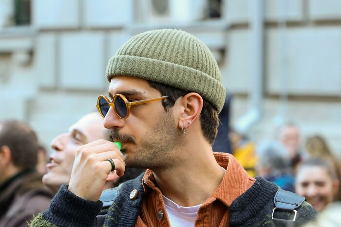 Man in sunglasses and a beanie sharing sounds in a crowd with others looking extremely angry around him.