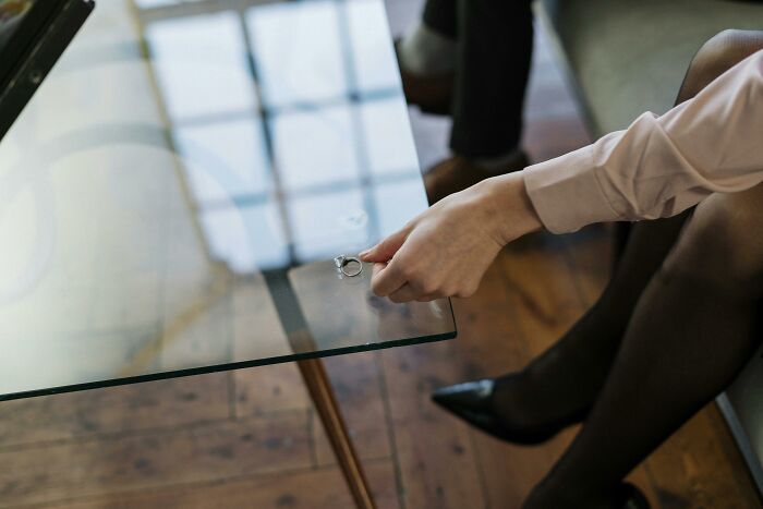 Woman wearing black stockings and pointed shoes holding a wedding ring near a glass table symbolizing marriage pressure and sad ending updates.