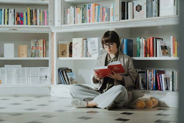 Young woman with glasses sitting on floor in library, reading a red book, showing attention to detail and spotting spelling mistakes.
