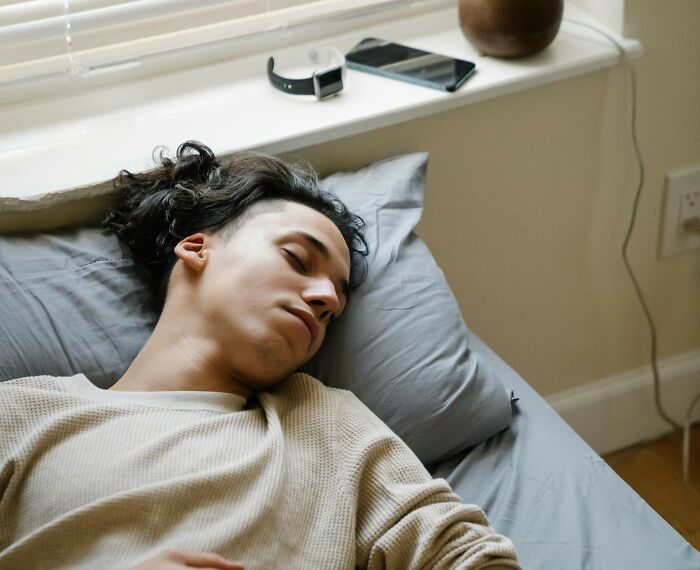 Young man sleeping on bed near window with smartphone and smartwatch, reflecting on nasty things exes did to partners.