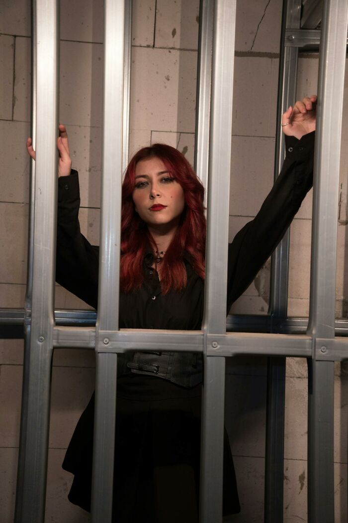 Woman with red hair wearing black behind metal bars, representing the villain in someone else’s story concept.