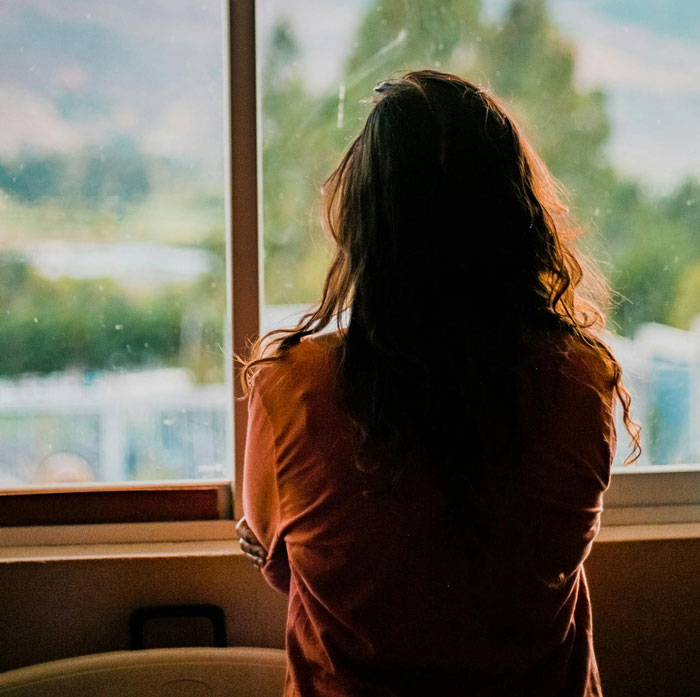 Person with long hair looking out a window, appearing thoughtful and worried about potential CPS call over dogs in house.