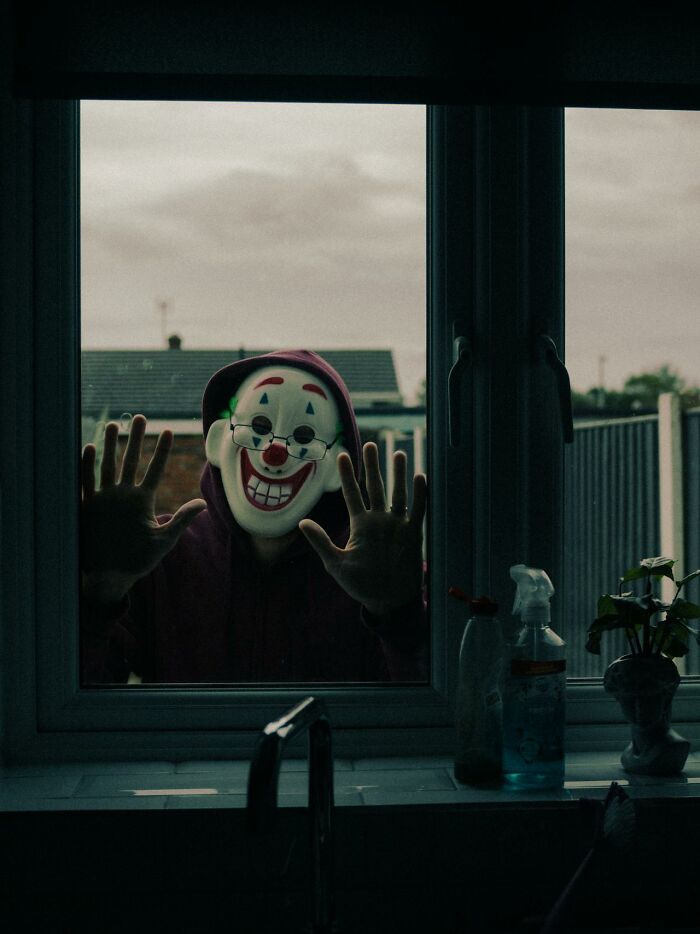 Person wearing a scary clown mask and hoodie outside a window, creating a horrifying home alone moment.