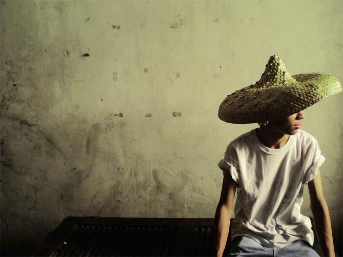 Young person wearing a large straw hat sitting against a worn wall, evoking a creepy stories atmosphere in dark woods.
