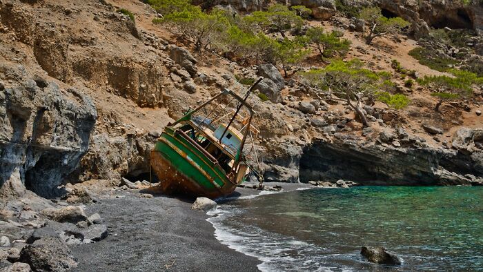 Abandoned boat on rocky shore near clear water with rugged cliffs and sparse trees in the background.