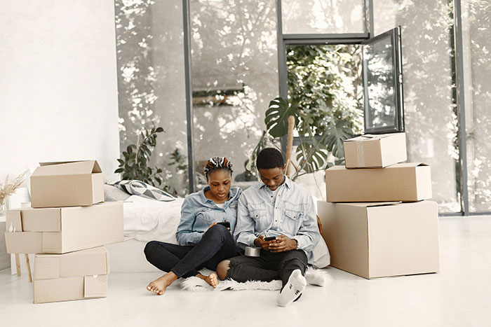 Couple sitting on floor surrounded by moving boxes, discussing memory box dilemma involving ex-girlfriend and late ex-boyfriend items.