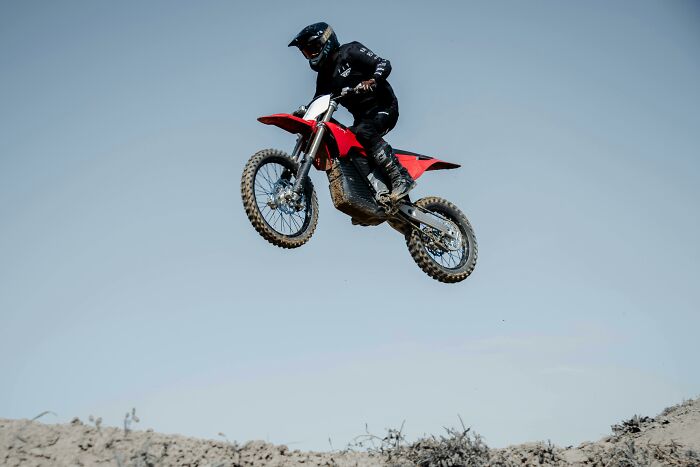 Person wearing black gear jumping a red dirt bike, with people sharing sounds of extreme anger implied in the scene.