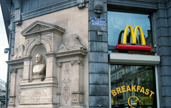 McDonald's storefront with breakfast sign next to historic stone monument in urban city setting overhyped products.