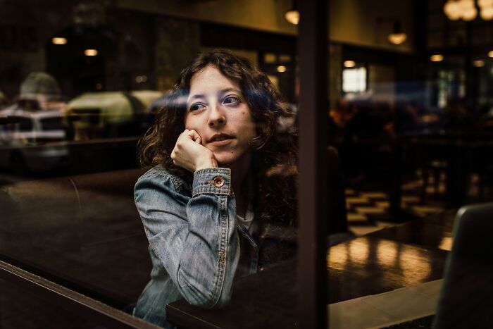 Young woman in a denim jacket sitting indoors, looking thoughtfully out a window, reflecting on pointless time spent.
