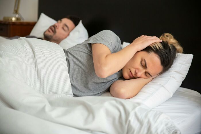 Couple in bed, woman covering ears in frustration while man sleeps, illustrating people share sounds extremely angry.