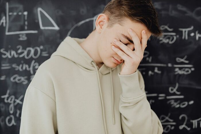 Teen boy in beige hoodie covering face in disbelief in front of chalkboard filled with complex math equations, showing unbelievable moments