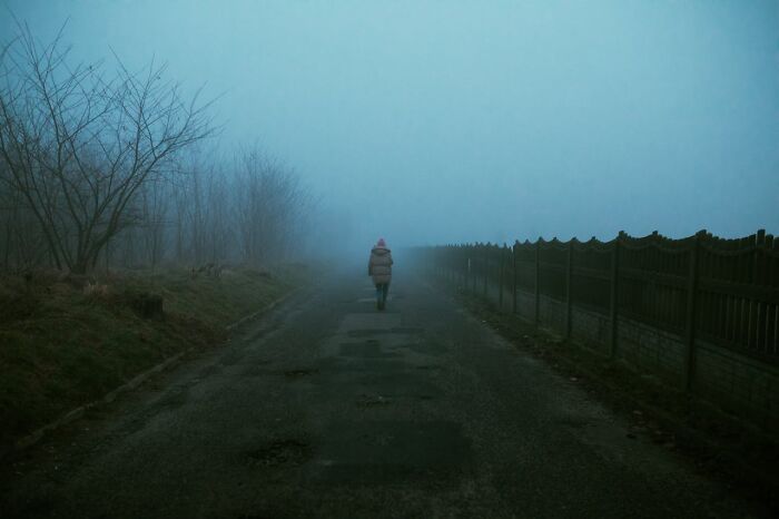 Person walking alone on a foggy road beside a fence and barren trees, capturing the creepy woods solitude experience.