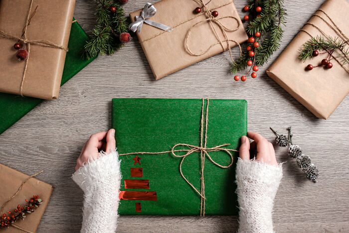 Hands holding a green wrapped gift surrounded by other rustic holiday presents and festive pine decorations
