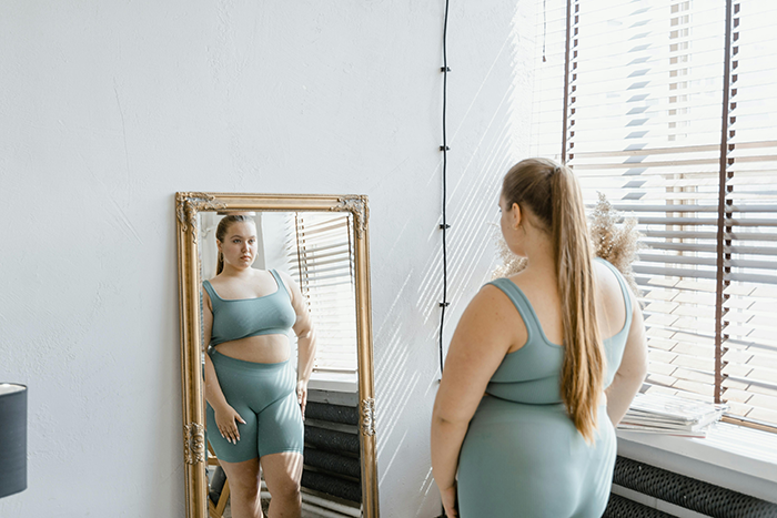 Woman in green workout clothes looks at herself in mirror, reflecting themes of shallow judgment and weight changes.