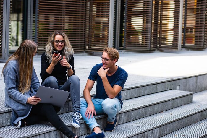 Three young people sitting on urban stairs with laptop and phone, illustrating people who were the villain in someone else’s story.