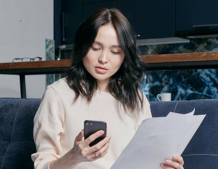 Young woman sitting on a couch looking shocked while reading paper and holding phone, reflecting nasty things exes did.