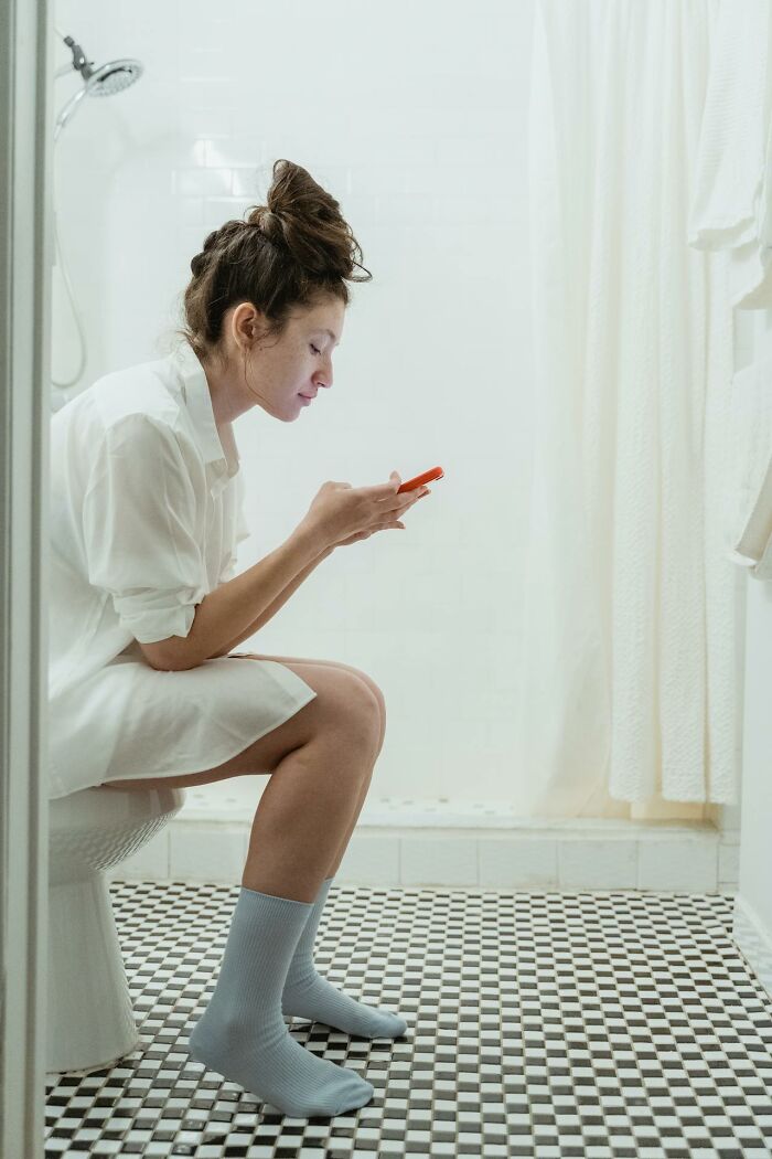 Woman sitting on toilet in bathroom looking at her phone, illustrating things women are not allowed to do freely.