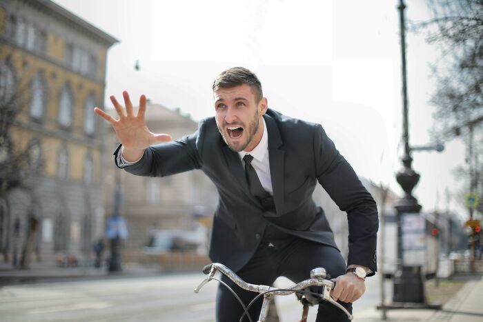 Man in a suit on a bicycle reaching out and sharing sounds while looking extremely angry on a city street.